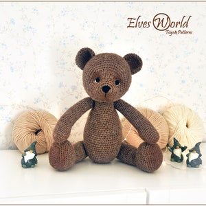 May include: A brown crocheted teddy bear sitting on a white surface. The bear has black eyes and a black nose. There are balls of yarn and small gnome figurines in the background.