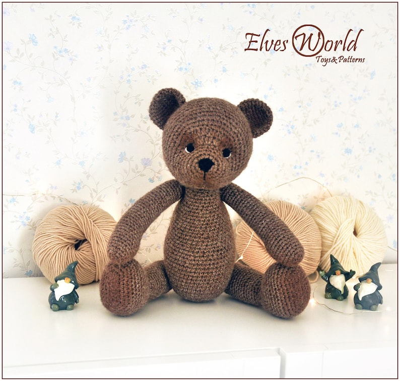 May include: A brown crocheted teddy bear sitting on a white surface. The bear has black eyes and a black nose. There are balls of yarn and small gnome figurines in the background.