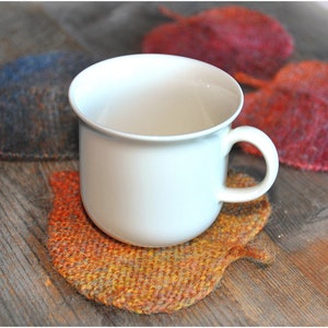 K&ouml;nnte beinhalten: Eine wei&szlig;e Keramiktasse steht auf einem gestrickten Untersetzer in Form eines Blattes. Der Untersetzer ist in Orange-, Gelb- und Braunt&ouml;nen gehalten.