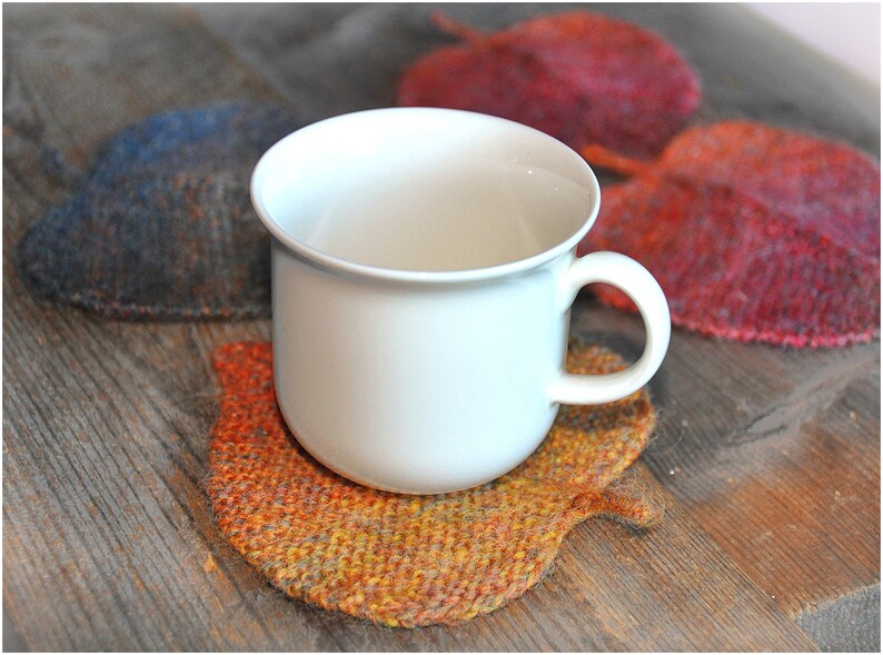 K&ouml;nnte beinhalten: Eine wei&szlig;e Keramiktasse steht auf einem gestrickten Untersetzer in Form eines Blattes. Der Untersetzer ist in Orange-, Gelb- und Braunt&ouml;nen gehalten.