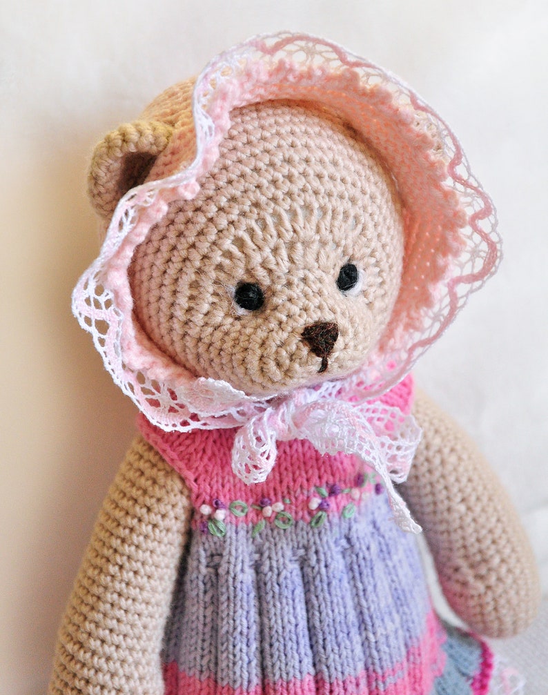 May include: A crocheted teddy bear wearing a pink and white dress with a lace trim. The bear has brown eyes and a brown nose.