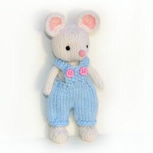 K&ouml;nnte beinhalten: Eine wei&szlig;e gestrickte Maus, die eine blaue Latzhose mit rosa Kn&ouml;pfen tr&auml;gt. Die Maus hat rosa Ohren und eine rosa Nase.