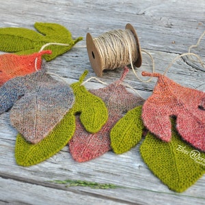 Puede incluir: Una colección de adornos de hojas de punto en tonos de verde, rojo y marrón, ensartados con un cordel. Las hojas están dispuestas en semicírculo sobre una superficie de madera. Un carrete de cordel es visible en el fondo.