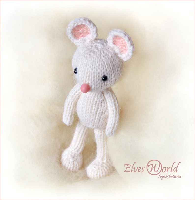K&ouml;nnte beinhalten: Ein wei&szlig;es, gestricktes Mausschmusetier mit rosa Ohren und einer rosa Nase. Das Spielzeug steht auf einer wei&szlig;en, flauschigen Oberfl&auml;che. Der Text "Elves World Toys & Patterns" ist in der rechten unteren Ecke des Bildes zu sehen.
