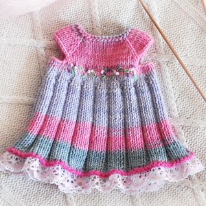 May include: A knitted baby dress with a purple, pink, and gray striped pattern. The dress has a white lace trim and a floral embroidery detail.