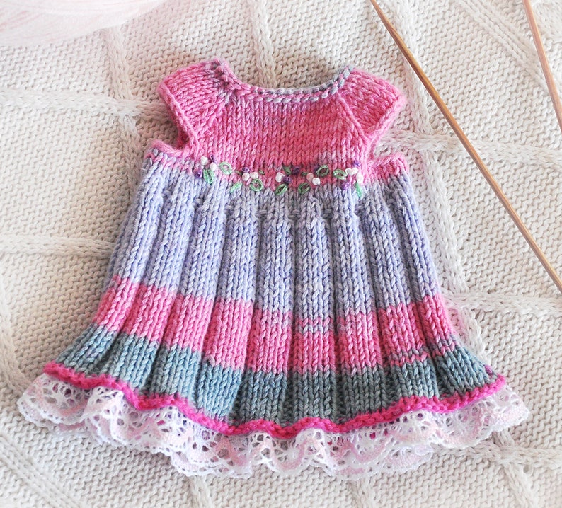 May include: A knitted baby dress with a purple, pink, and gray striped pattern. The dress has a white lace trim and a floral embroidery detail.
