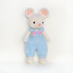 K&ouml;nnte beinhalten: Eine wei&szlig;e gestrickte Maus, die eine blaue Latzhose mit rosa Kn&ouml;pfen tr&auml;gt. Die Maus hat rosa Ohren und eine rosa Nase.