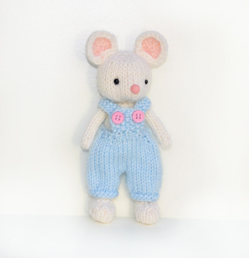 K&ouml;nnte beinhalten: Eine wei&szlig;e gestrickte Maus, die eine blaue Latzhose mit rosa Kn&ouml;pfen tr&auml;gt. Die Maus hat rosa Ohren und eine rosa Nase.