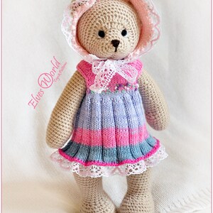 May include: A crocheted teddy bear wearing a pink and white dress with a lace trim. The bear is wearing a pink and white crocheted bonnet with a lace trim. The bear is standing on a white surface.