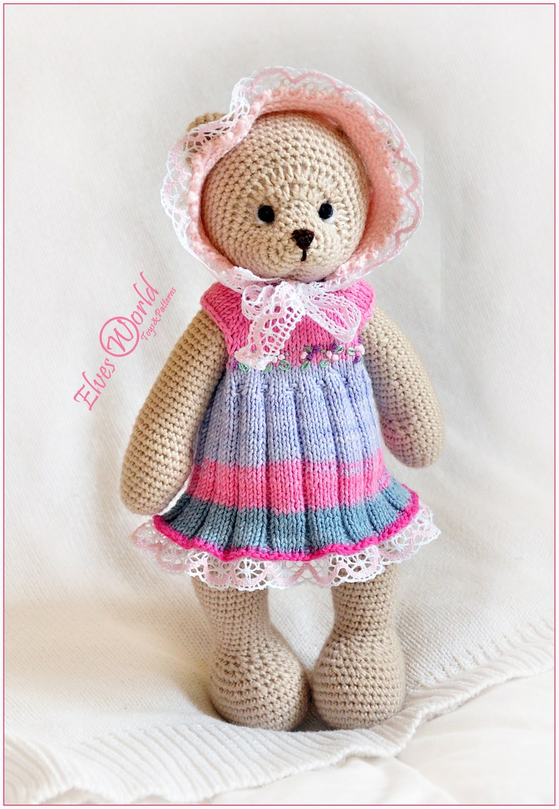 May include: A crocheted teddy bear wearing a pink and white dress with a lace trim. The bear is wearing a pink and white crocheted bonnet with a lace trim. The bear is standing on a white surface.