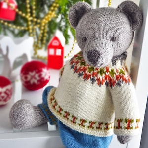 Puede incluir: Un oso de peluche gris tejido, con un suéter crema con un patrón geométrico colorido y pantalones azules. El oso tiene una nariz y orejas de color gris oscuro. Decoraciones navideñas en el fondo.