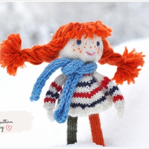 Puede incluir: Muñeca de punto con pelo rojo, una bufanda azul y un suéter a rayas. La muñeca está de pie en la nieve. El texto "PDF pattern knitting" es visible en la esquina inferior izquierda de la imagen.