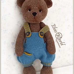 May include: A brown crocheted teddy bear wearing blue dungarees with yellow accents. The bear has a black nose and eyes. The text "Elves World Toys & Patterns" is visible in the background.