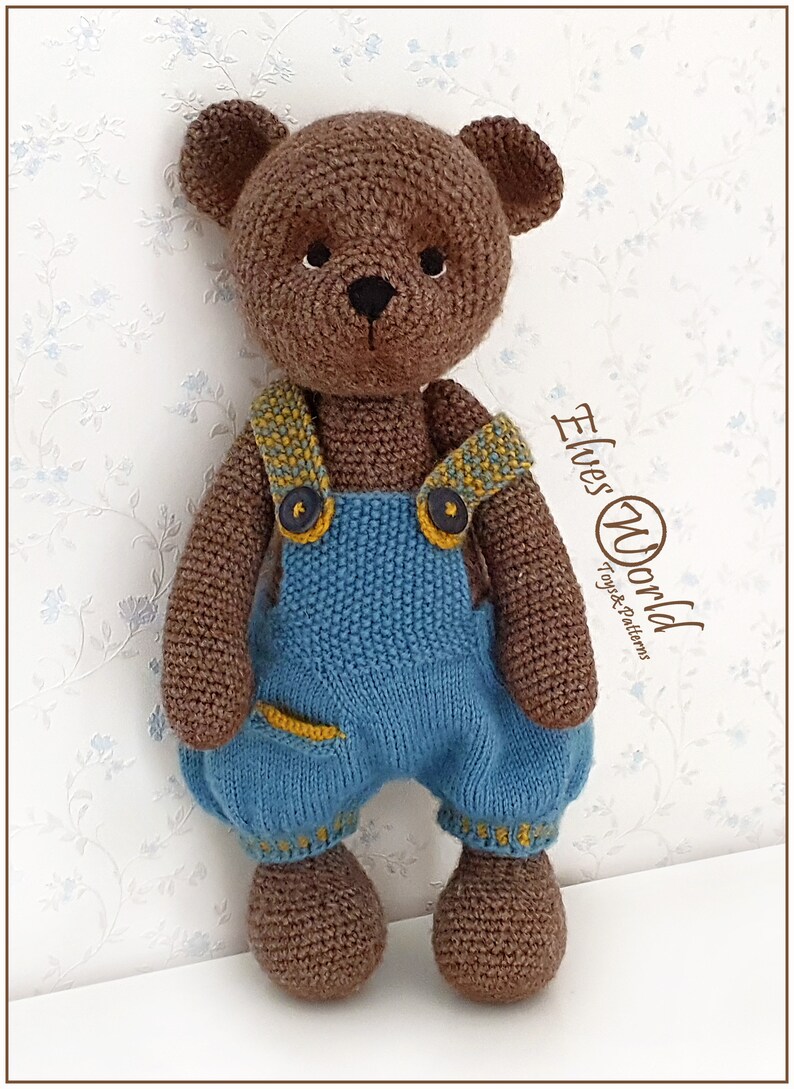 May include: A brown crocheted teddy bear wearing blue dungarees with yellow accents. The bear has a black nose and eyes. The text "Elves World Toys & Patterns" is visible in the background.