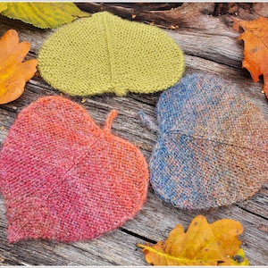 K&ouml;nnte beinhalten: Drei gestrickte Untersetzer in Blattform in verschiedenen Farben: gr&uuml;n, rosa und blau. Die Untersetzer sind auf einer Holzoberfl&auml;che mit Herbstbl&auml;ttern angeordnet.