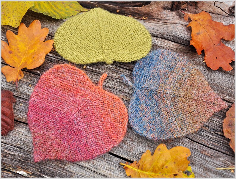 K&ouml;nnte beinhalten: Drei gestrickte Untersetzer in Blattform in verschiedenen Farben: gr&uuml;n, rosa und blau. Die Untersetzer sind auf einer Holzoberfl&auml;che mit Herbstbl&auml;ttern angeordnet.