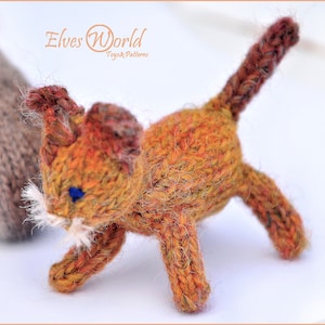 Puede incluir: Un juguete de gato de punto naranja y marrón con ojos azules y bigotes blancos. El juguete está hecho por Elves World Toys & Patterns.