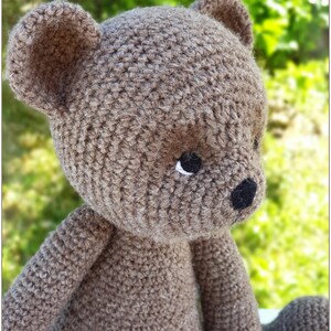 May include: A crocheted brown teddy bear with black eyes and a black nose. The bear is sitting on a green and white background.