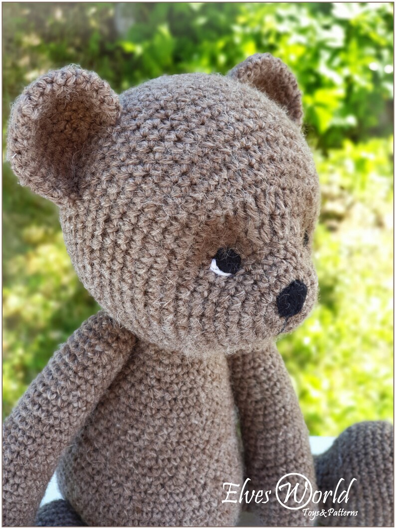 May include: A crocheted brown teddy bear with black eyes and a black nose. The bear is sitting on a green and white background.