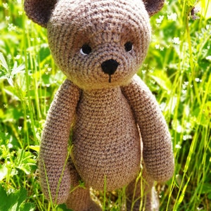 May include: A brown crocheted teddy bear standing in a field of green grass with white flowers.