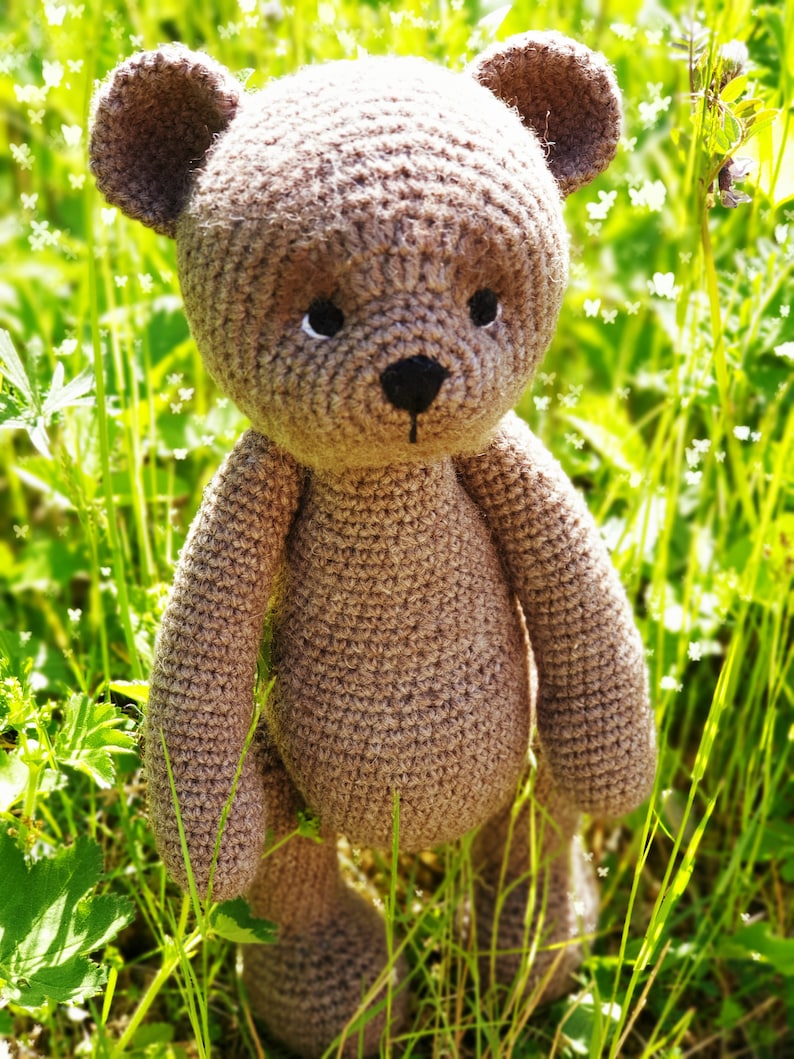 May include: A brown crocheted teddy bear standing in a field of green grass with white flowers.