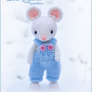 K&ouml;nnte beinhalten: Eine wei&szlig;e gestrickte Maus, die eine blaue Latzhose mit rosa Kn&ouml;pfen tr&auml;gt. Die Maus hat rosa Ohren und eine rosa Nase. Die Maus steht auf einer wei&szlig;en Oberfl&auml;che.