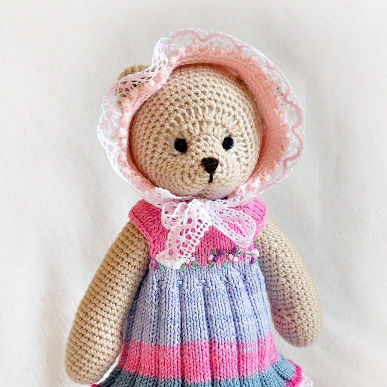 May include: A crocheted teddy bear wearing a pink and white bonnet and a lavender and pink striped dress.