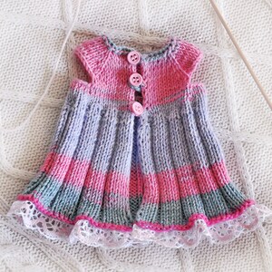 May include: A knitted baby dress with pink, purple, and gray stripes. The dress has a lace trim and pink buttons down the front.