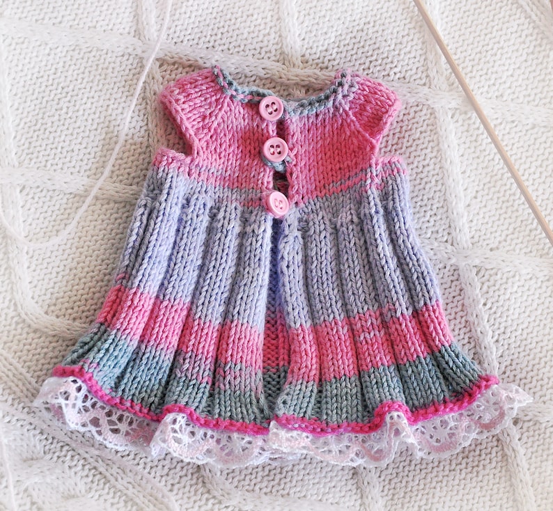 May include: A knitted baby dress with pink, purple, and gray stripes. The dress has a lace trim and pink buttons down the front.