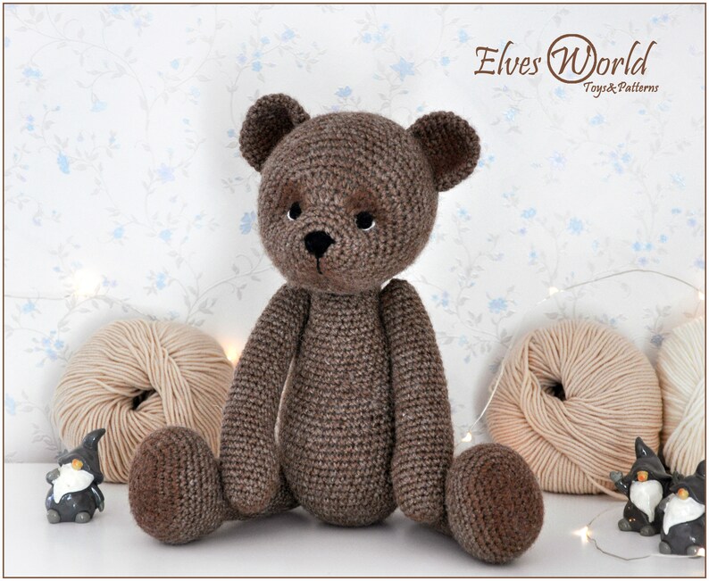 May include: A brown crocheted teddy bear sitting on a white surface with two balls of yarn and two small ceramic figurines. The bear has black eyes and a black nose.