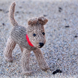 Könnte beinhalten: Ein braunes, gestricktes Hundespielzeug mit einem roten Band um den Hals. Das Spielzeug liegt auf einer grauen und braunen, strukturierten Oberfläche.