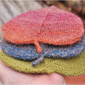 K&ouml;nnte beinhalten: Drei gestrickte, blattf&ouml;rmige M&uuml;tzen, die &uuml;bereinander gestapelt sind. Die oberste M&uuml;tze ist rot-orange, die mittlere M&uuml;tze ist blau-grau und die unterste M&uuml;tze ist gr&uuml;n.