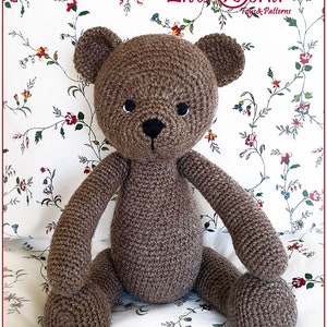 May include: A brown crocheted teddy bear sitting on a white floral patterned fabric. The bear has black eyes and a black nose.