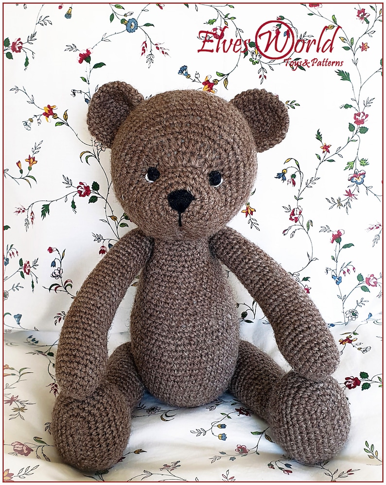 May include: A brown crocheted teddy bear sitting on a white floral patterned fabric. The bear has black eyes and a black nose.