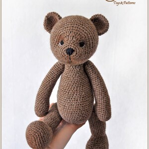 May include: A brown crocheted teddy bear with black eyes and a black nose. The bear is sitting on a hand.