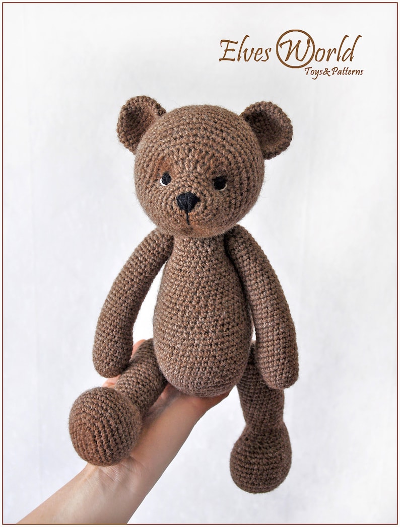 May include: A brown crocheted teddy bear with black eyes and a black nose. The bear is sitting on a hand.