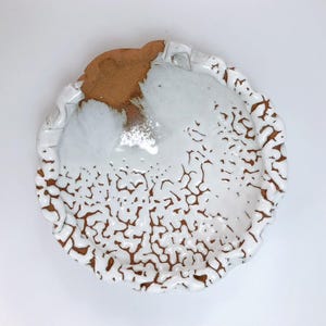 Puede incluir: Un plato de cerámica hecho a mano con un diseño único. El plato presenta una superficie esmaltada blanca con un patrón marrón agrietado en los bordes. El borde tiene un diseño texturizado y ondulado. El plato tiene aproximadamente 20 cm de diámetro.