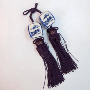 May include: Two decorative tassels with ceramic square pendants. The pendants feature a blue and white landscape design. The tassels are a deep purple colour, with a knotted top and long, flowing strands.