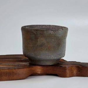 Puede incluir: Taza de cerámica artesanal con una paleta de colores oscuros y terrosos. La taza tiene una forma ligeramente irregular y una superficie texturizada, lo que sugiere una estética natural y orgánica. Se apoya sobre una base de madera oscura, acentuando su aspecto artesanal.