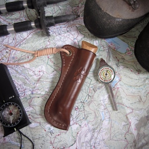 Op de afbeelding: Een bruine leren schede met een mes met houten handvat erin. De schede heeft een bruine leren riem met een metalen gesp. Het mes ligt op een kaart met een kompas en wandelstokken.