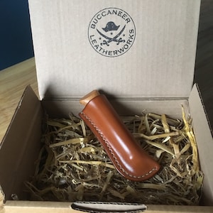 Op de afbeelding: Een bruine leren mes schede met een houten handvat, in een kartonnen doos met verpakkingsmateriaal. De doos heeft een zwart-wit logo met de tekst "Buccaneer Leatherworks" met een piratenhoed en gekruiste zwaarden.