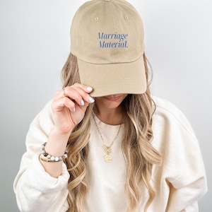 May include: A tan baseball cap with the text "Marriage Material." embroidered in blue on the front.