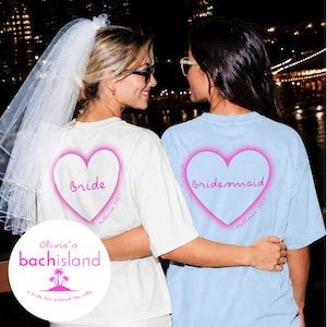 May include: A white t-shirt with a pink heart graphic reading Bride and a light blue t-shirt with a pink heart graphic reading bridesmaid, both featuring the text Mallorca 2027. A white veil is attached to the white t-shirt.