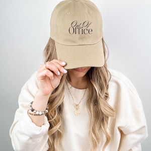 Może przedstawiać: Beżowa czapka z daszkiem z czarnym napisem "Out Of Office". Czapka ma zakrzywiony daszek i jest wykonana z miękkiego materiału. Czapka jest trzymana przez osobę.