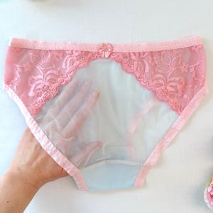 Könnte beinhalten: Hellrosa Slip mit einem transparenten Mittelteil und dekorativen floralen Spitzenverzierungen entlang des oberen Bundes. Eine kleine, passende rosa Schleife ist mittig am oberen Rand des Bundes angebracht und setzt einen dezenten Akzent.