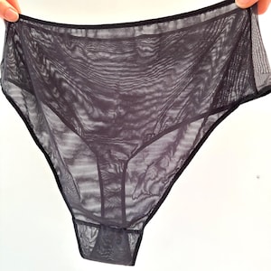 Op de afbeelding: Donkergrijze, doorschijnende onderbroek met een hoge taille. Het kledingstuk is gemaakt van een lichte, doorschijnende stof met zwarte randen. De onderbroek wordt door een hand omhoog gehouden, wat de delicate constructie en vorm laat zien.