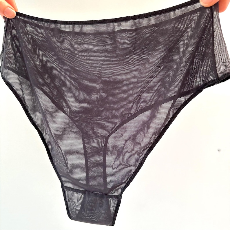 Op de afbeelding: Donkergrijze, doorschijnende onderbroek met een hoge taille. Het kledingstuk is gemaakt van een lichte, doorschijnende stof met zwarte randen. De onderbroek wordt door een hand omhoog gehouden, wat de delicate constructie en vorm laat zien.