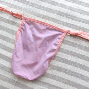 Könnte beinhalten: Ein helllila String mit pfirsichfarbener Bordüre und Trägern. Die Kanten sind mit einer dekorativen Naht versehen. Die Träger sind mit kleinen Metallringen verstellbar. Der String liegt auf einem gestreiften Stoff.