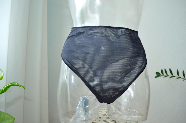 Op de afbeelding: Donker marineblauwe slip met hoge taille, weergegeven op een doorzichtige mannequin. De transparante stof heeft een subtiel gestreept patroon. De achtergrond omvat een wit gordijn en een groene plant.