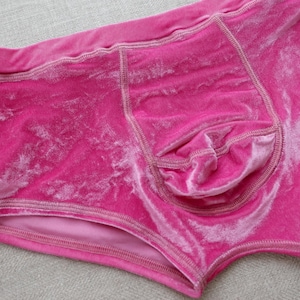Puede incluir: Un par de ropa interior de terciopelo rosa brillante con una textura suave y afelpada con un brillo sutil. La prenda incluye un diseño de bolsa frontal contorneada con costuras tonales visibles y una cintura elástica ancha.